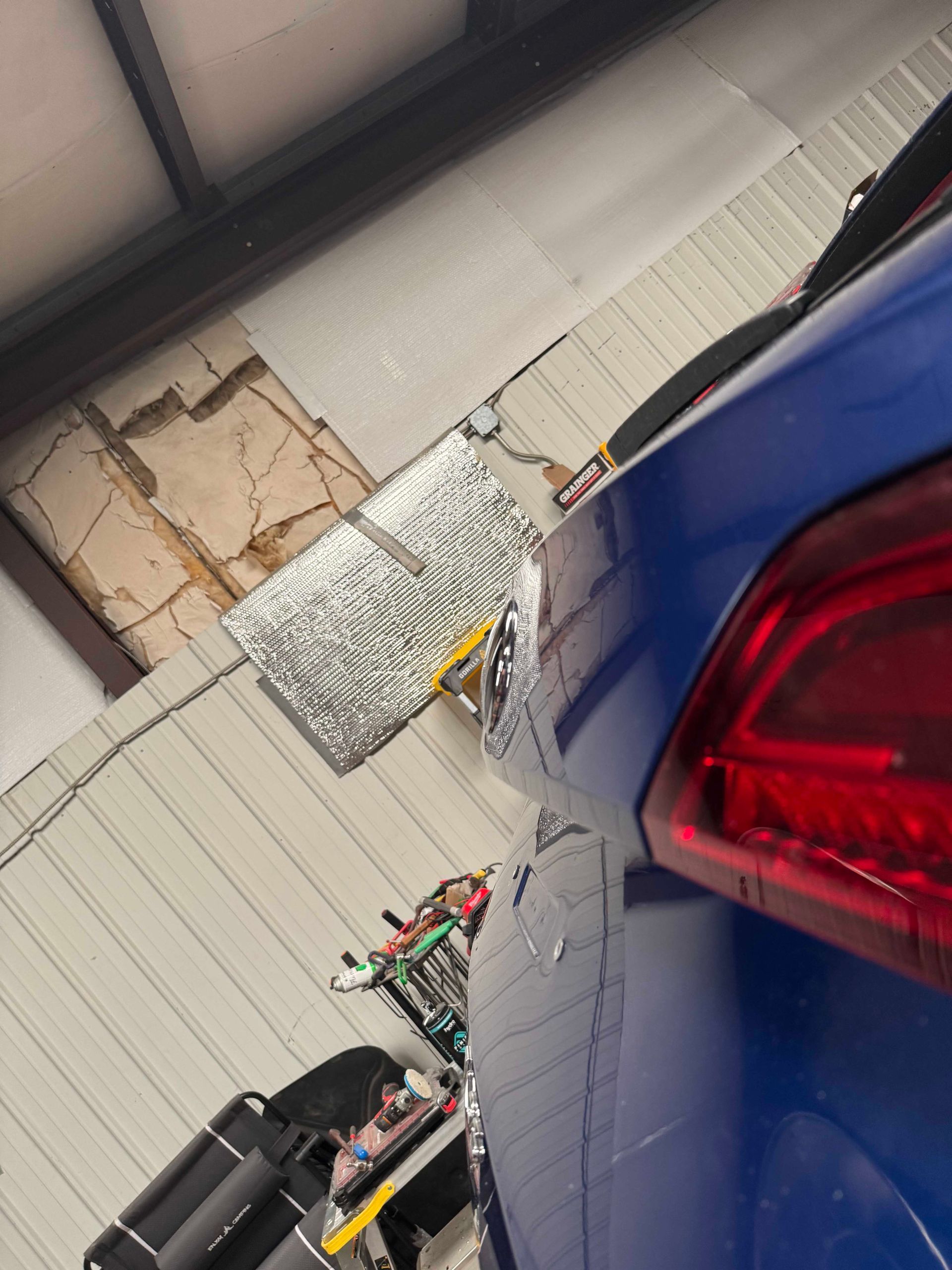 Blue car rear damaged, parked inside a garage. Damaged wall and tools visible.