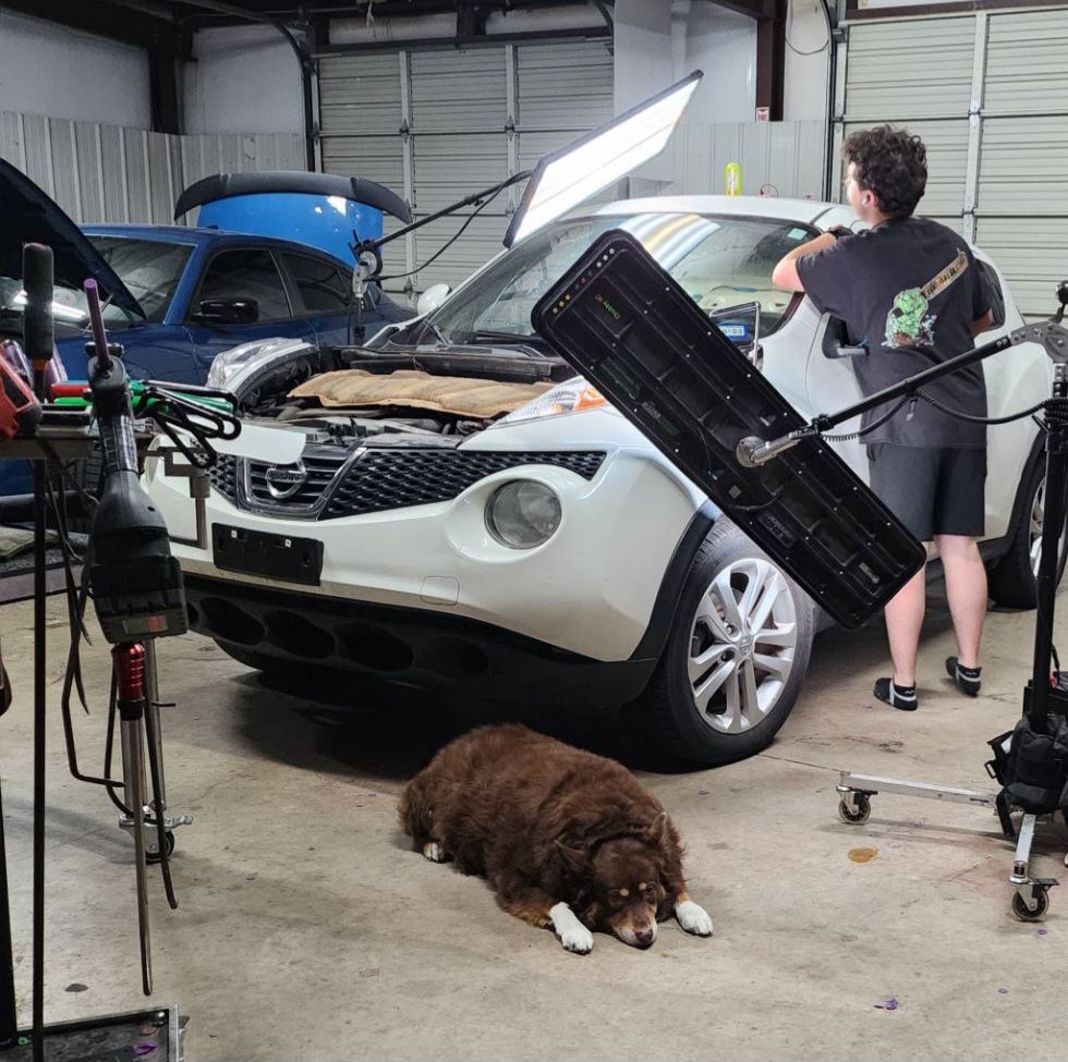 Man working on a white car in a garage with a dog lying on the ground. Lighting equipment set up.