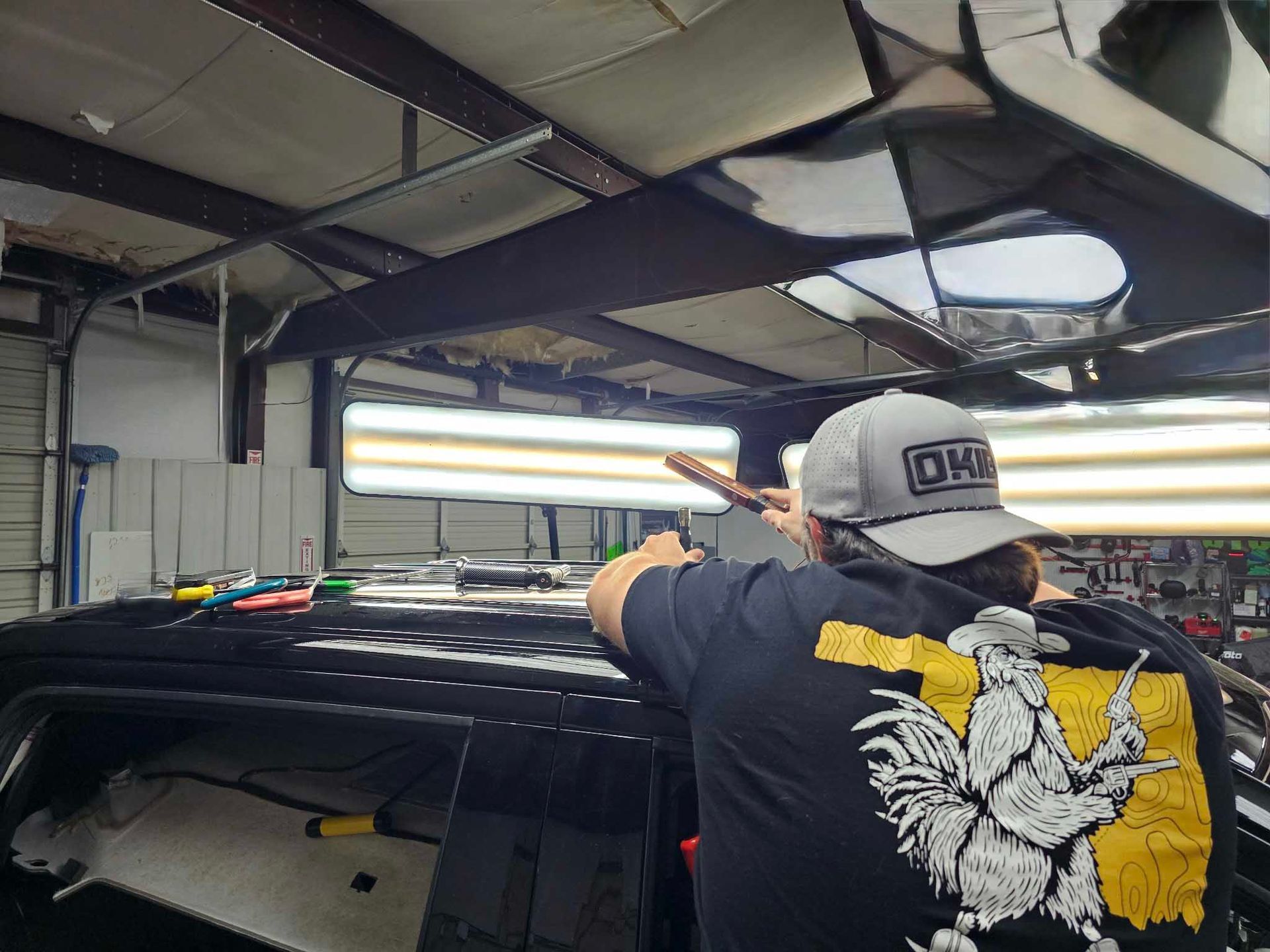 Person using tools to repair a dent on a black car roof inside a shop, with overhead lighting.