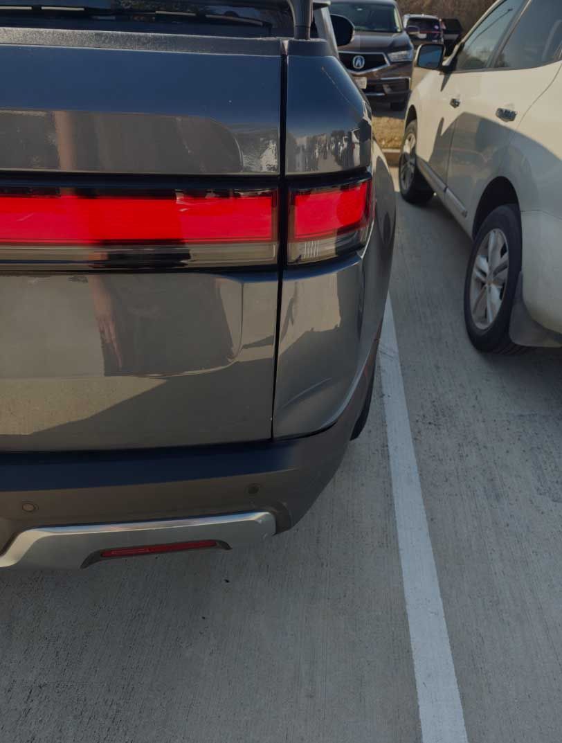 Rear view of a dark gray Rivian truck, next to a beige car in a parking spot. Red taillight.