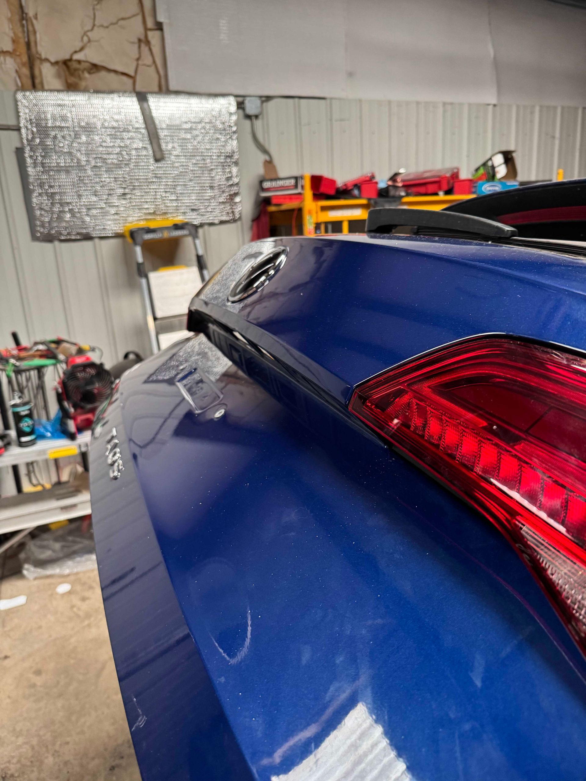 Blue car's rear, taillight visible. The car is inside a garage with reflective insulation on the wall.