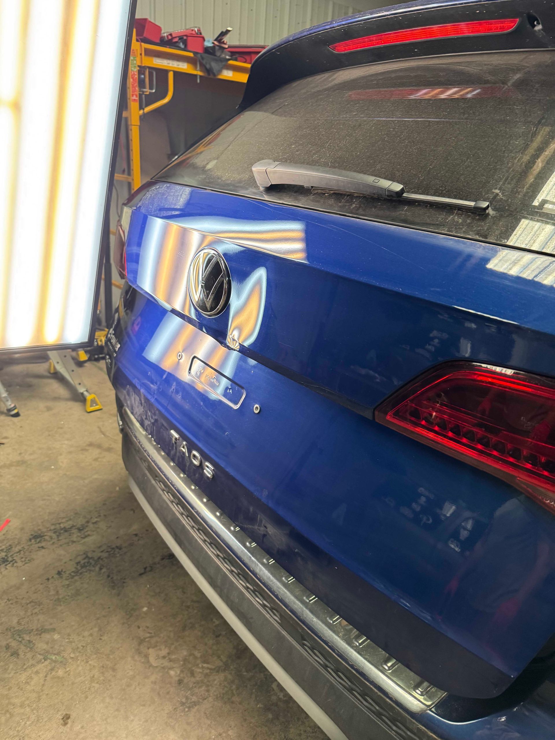 Blue car's rear with a dent, being worked on under a repair light.