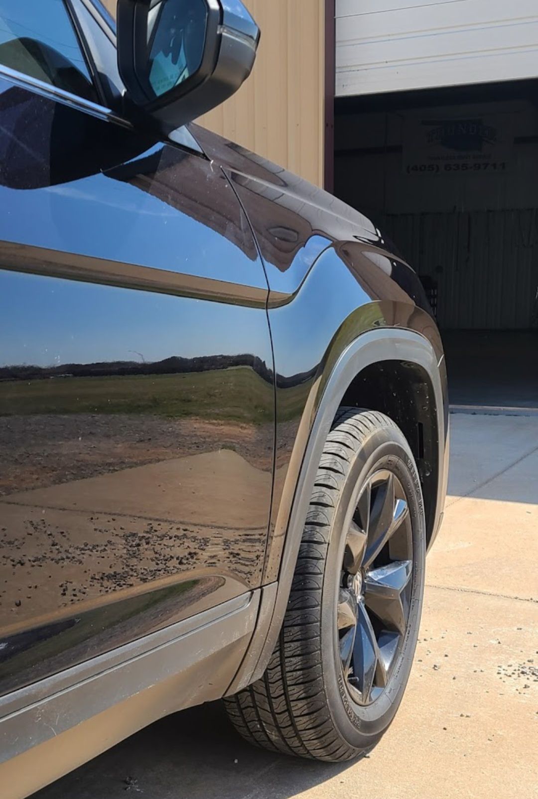 Black SUV parked near a garage with a view of a field and a partly cloudy sky reflected in the car's body.