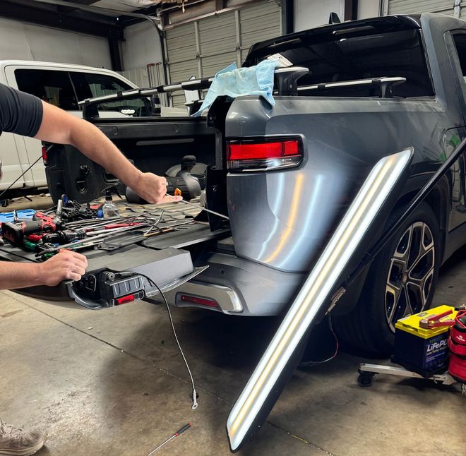 Person repairing dent on gray truck bed in a shop, using a light and tools.