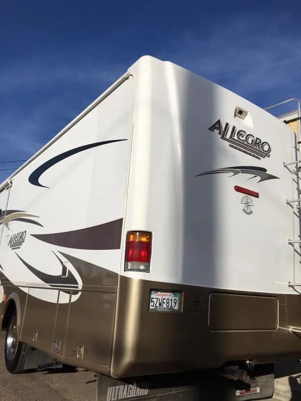 Rear view of a white and gold RV with "Allegro" logo. Features a ladder and taillight.