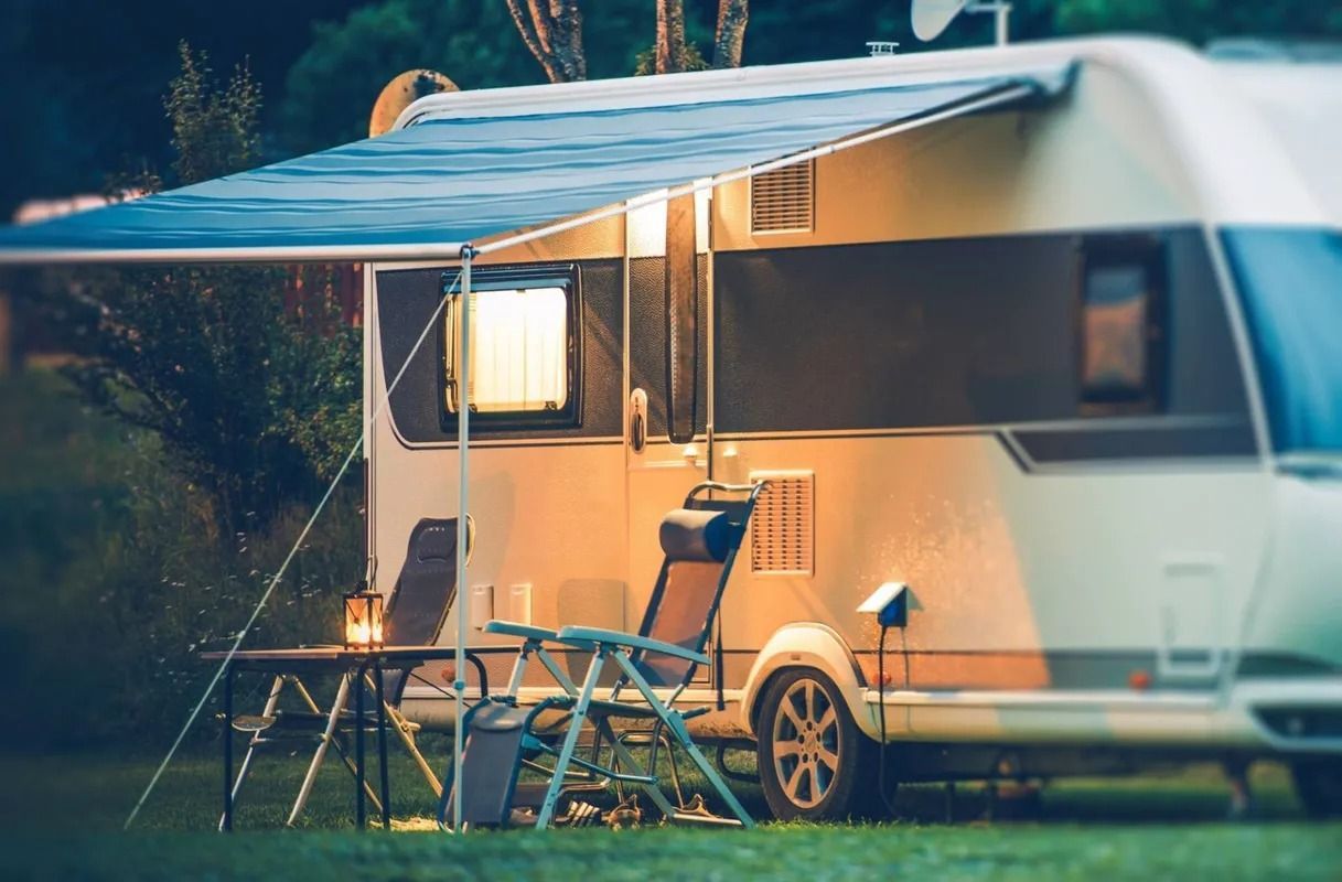 RV parked outdoors at night, awning extended, table and chairs set up. Warm light glows.