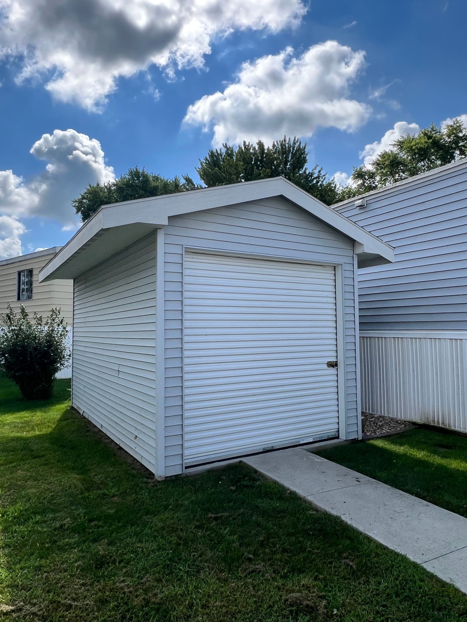 White garage with a rolling door, gray walkway, and grassy yard under a cloudy sky.