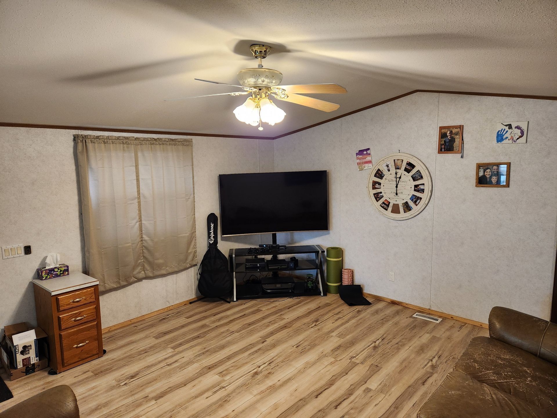 Living room with TV, guitar, clock, and tan ceiling fan. Wooden floor, light-colored walls, and tan curtains.