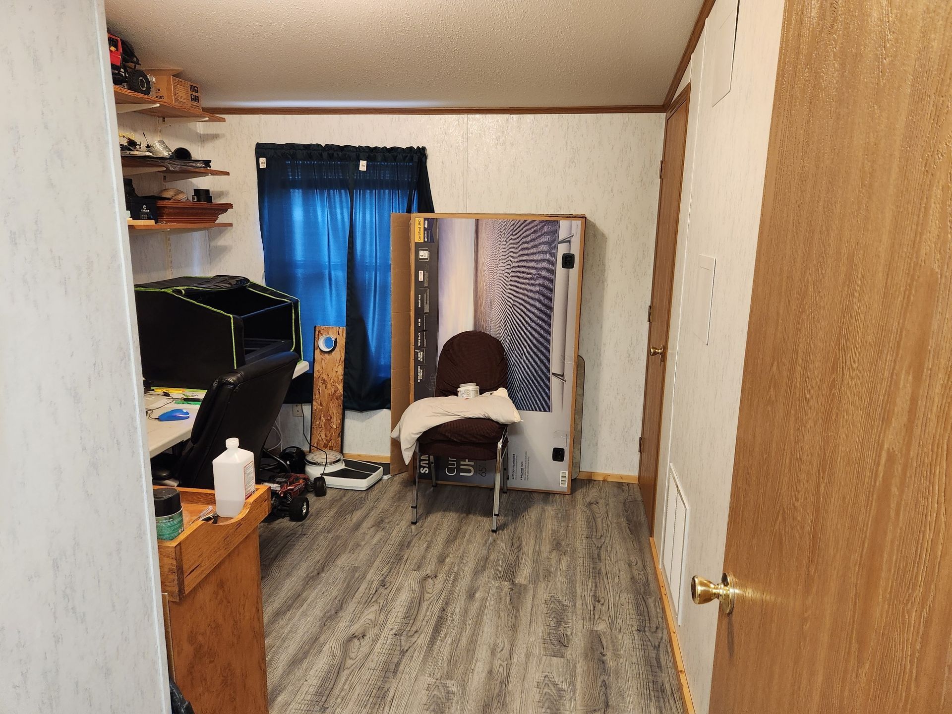 A cluttered room with a desk, chair, and large cardboard box near a window with blue curtains.