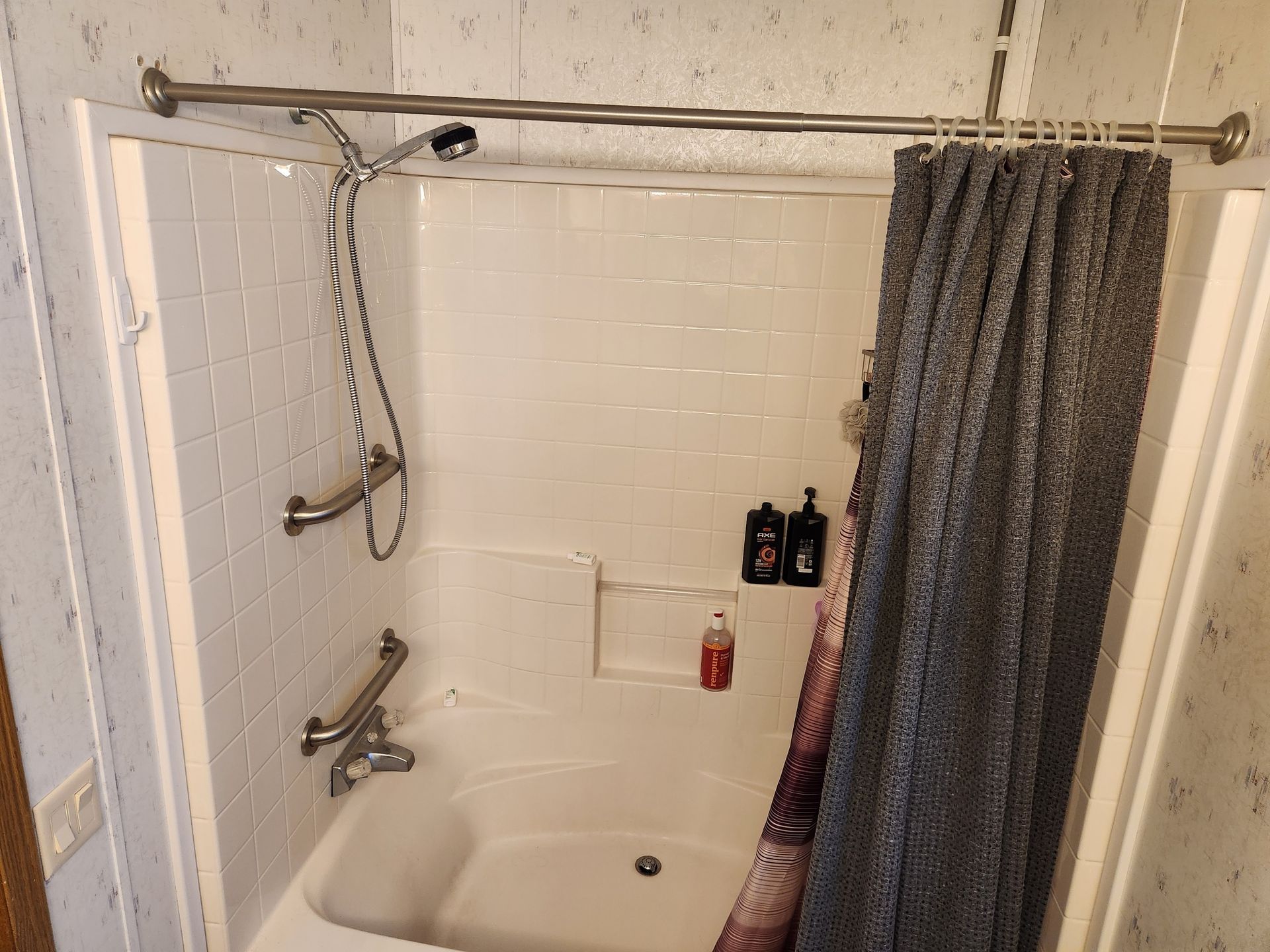 A small, white shower stall with a curtain. It has grab bars, soap bottles, and a hand-held shower head.