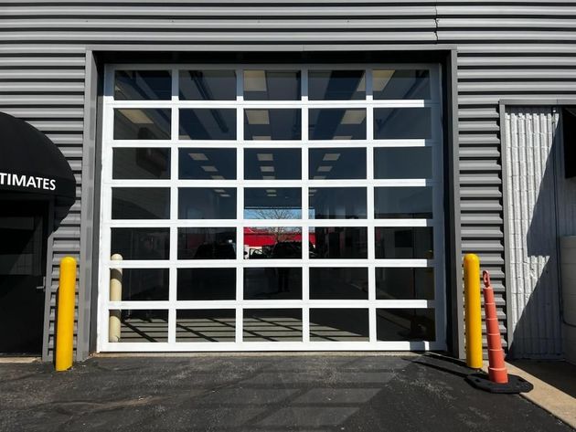 Garage door with white grid panels on a gray industrial building, flanked by yellow bollards and an orange cone