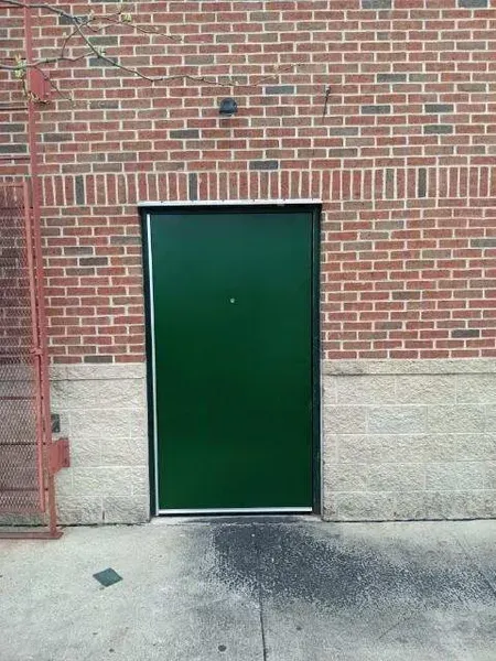 Green metal door in a brick wall with a concrete base and sidewalk in front