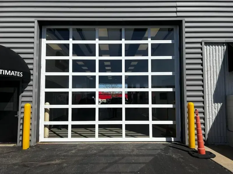 Glass garage door on a gray industrial building, with yellow bollards and a traffic cone nearby