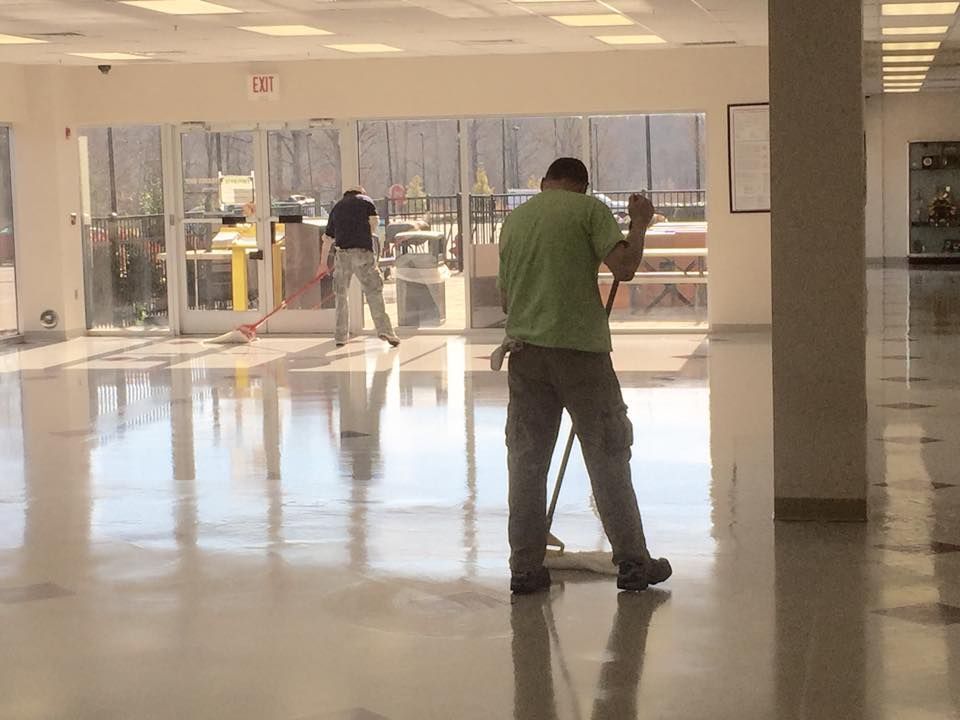 Two men mopping a shiny, light-colored floor inside a building with large windows.