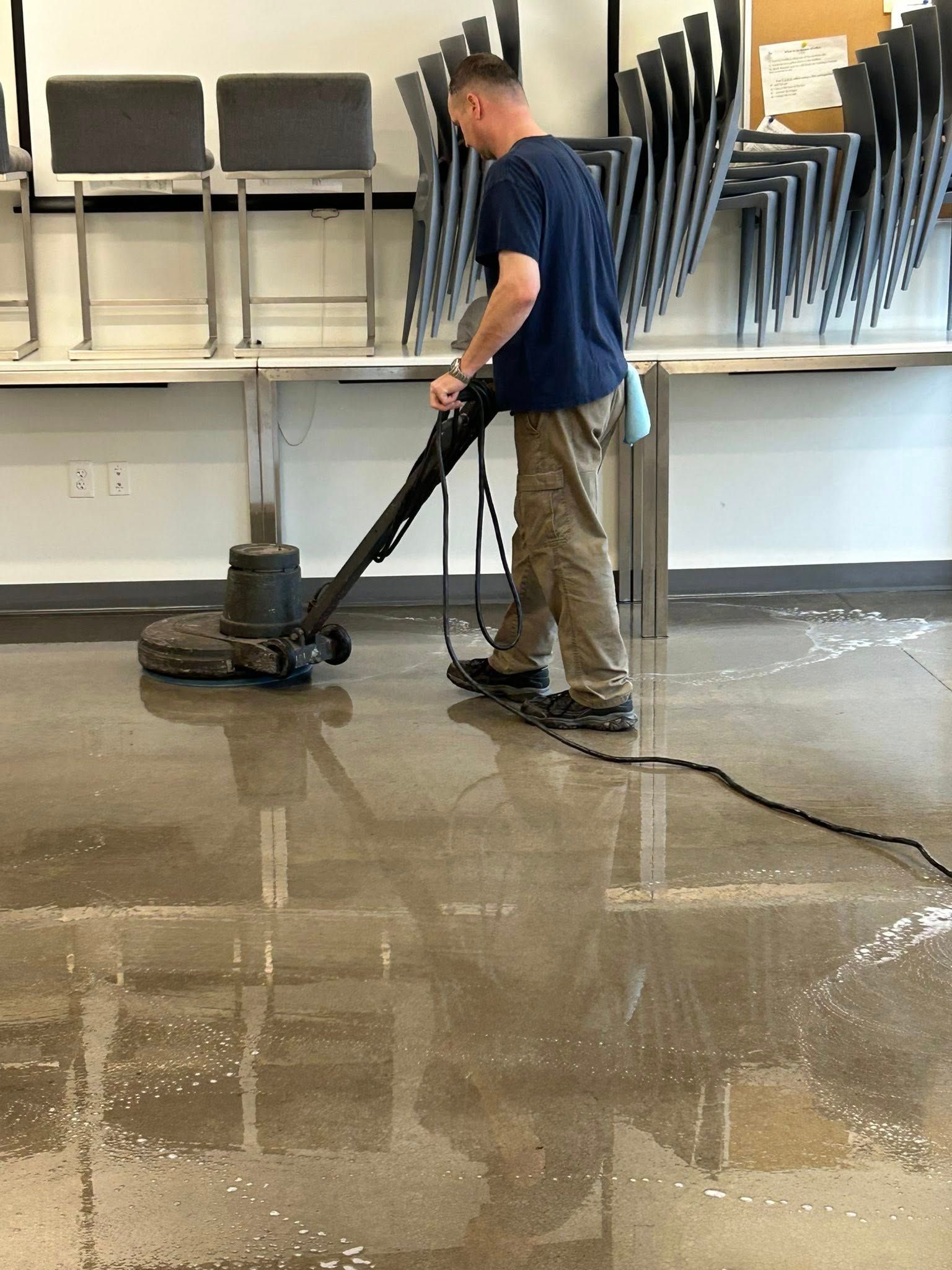 Man cleaning a concrete floor with a floor buffer in a room with tables and chairs.