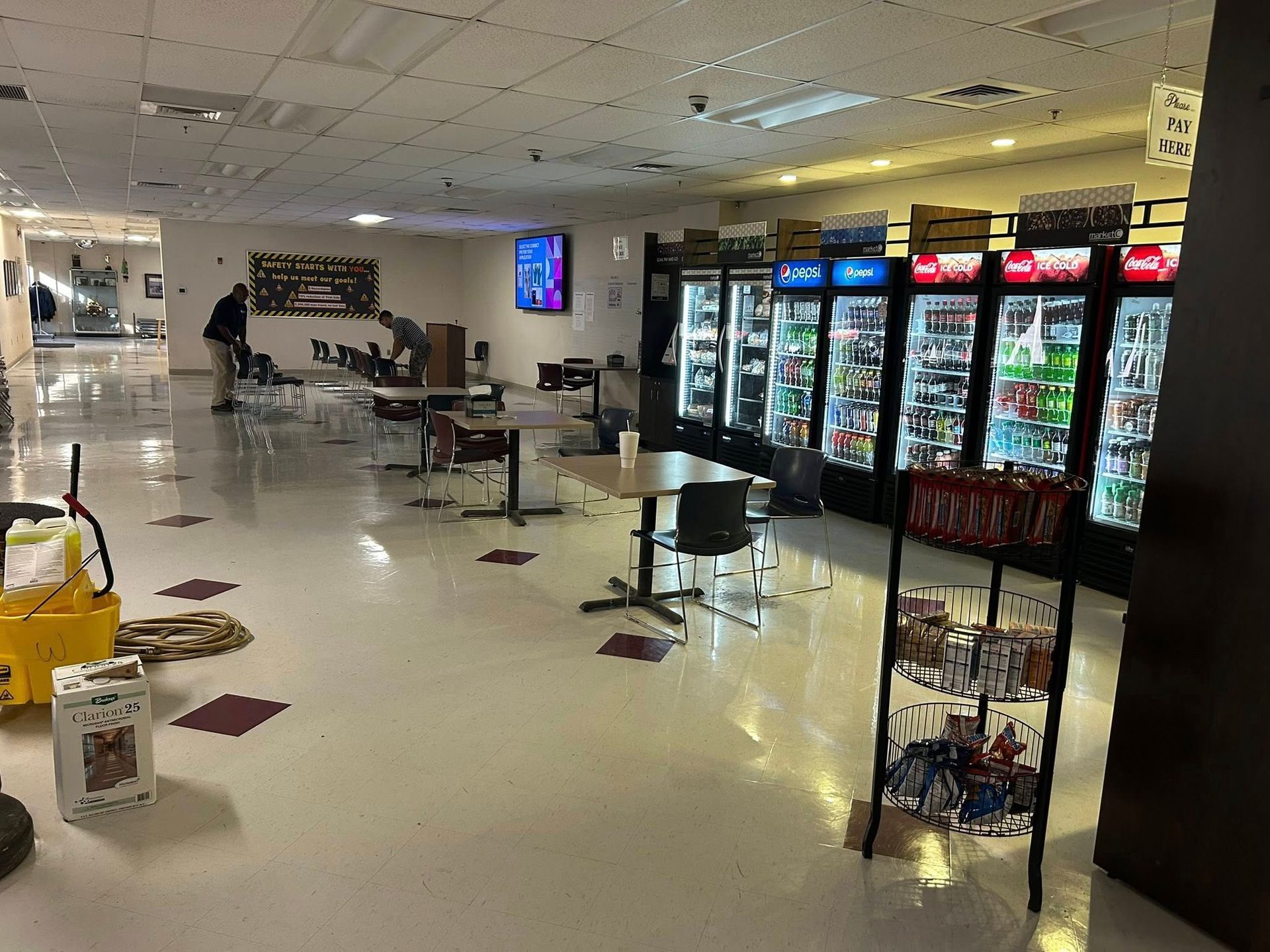 Interior view of a cafeteria/break area with vending machines, tables, and a person mopping the floor.
