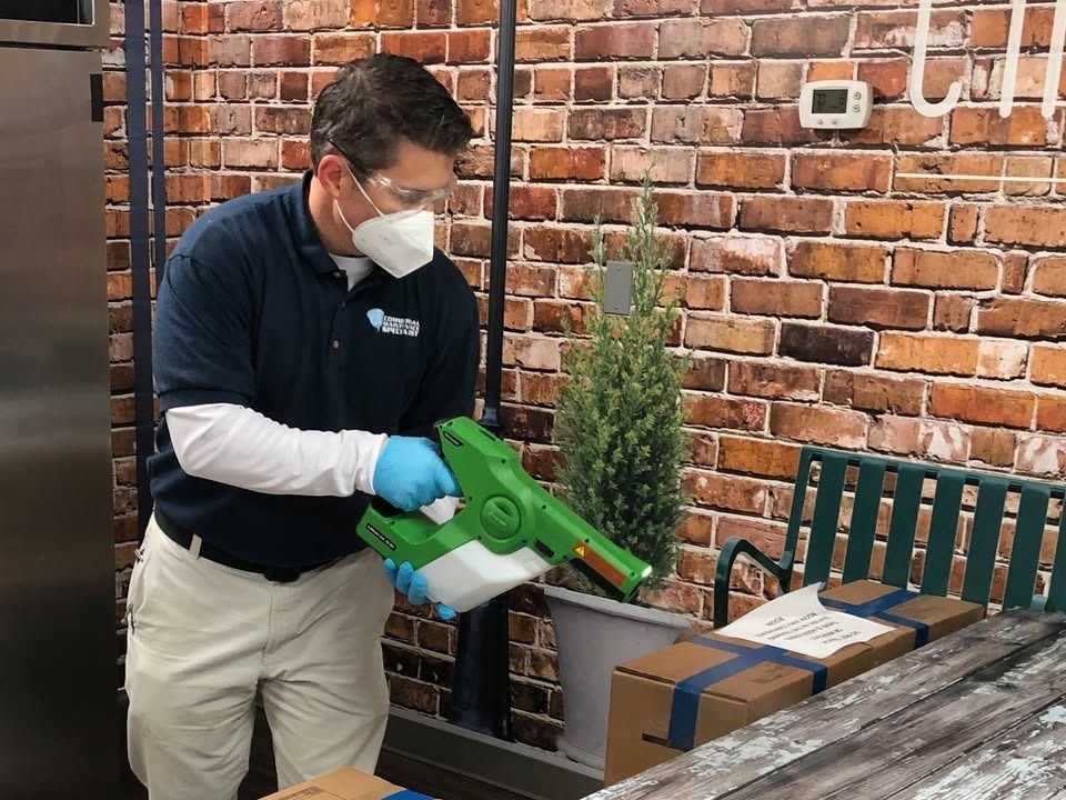 Man in mask and gloves disinfecting a table with a spray gun against a brick wall.