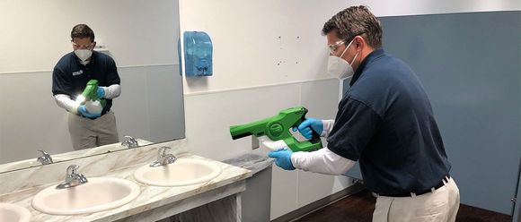 Man disinfecting a bathroom with a spray gun, wearing a mask, gloves, and safety glasses.