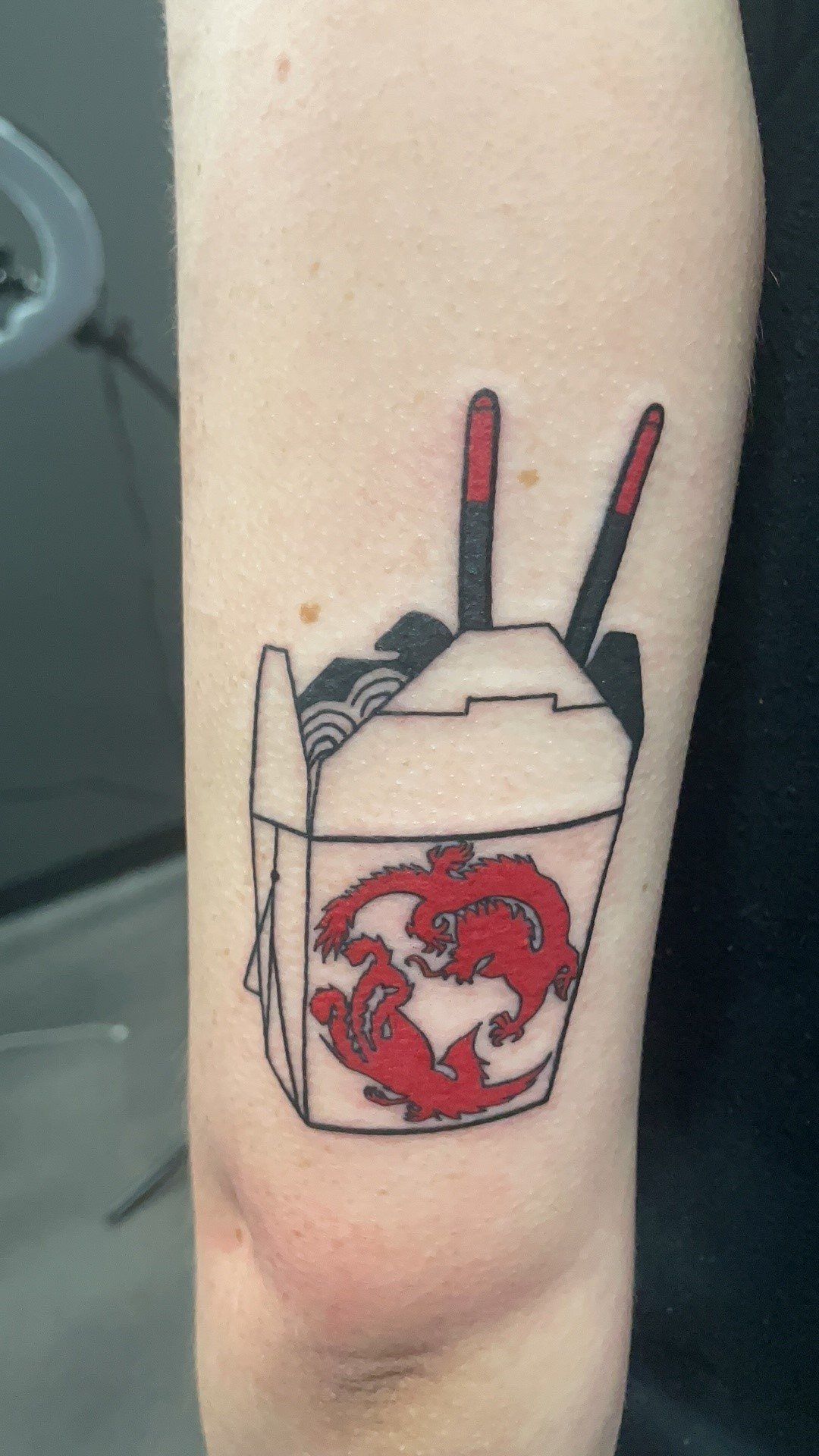 A person has a tattoo of a takeout box with chopsticks on their arm.