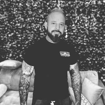 A man with a beard and tattoos is standing in front of a couch in a black and white photo.