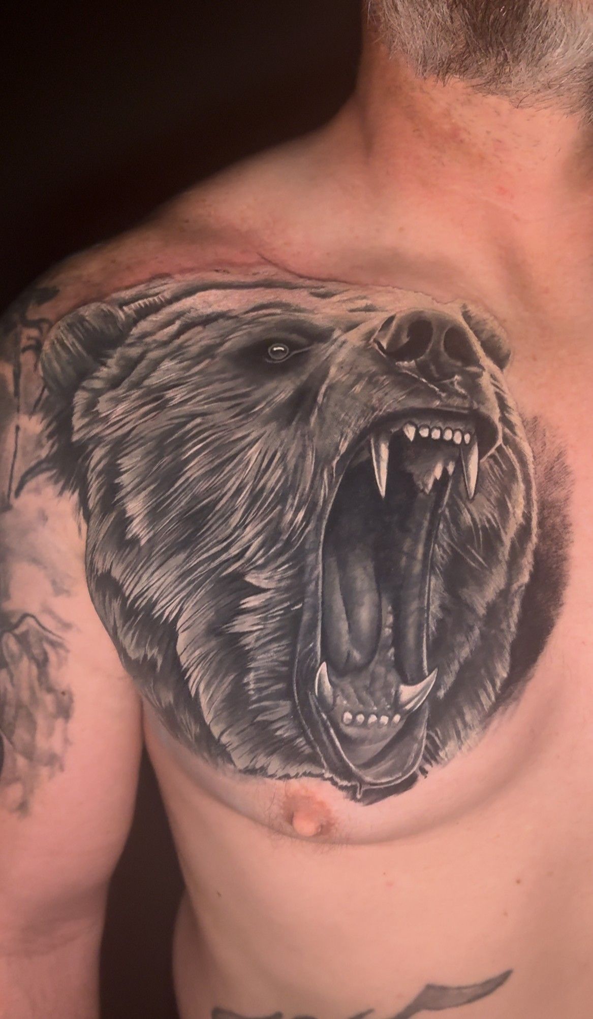 Man's chest tattoo: snarling bear head with detailed fur, open mouth, sharp teeth, dark background.