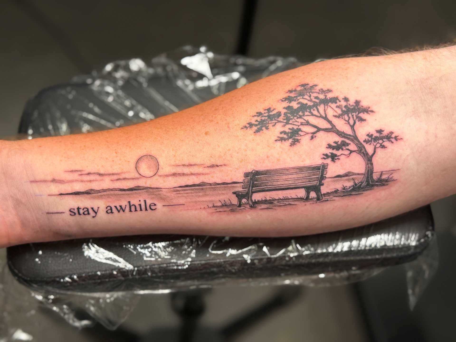 Forearm tattoo of a sunset bench scene with text and a tree silhouette