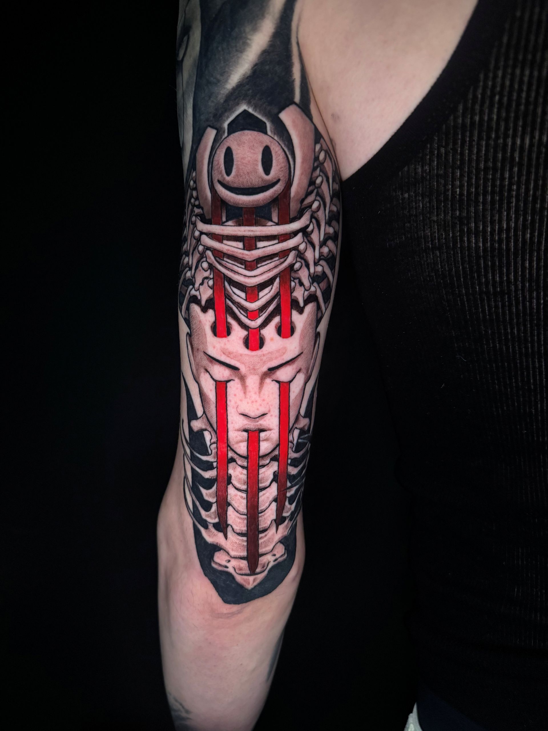 Abstract black and red forearm tattoo featuring a stylized smiling face enclosed within a skeletal ribcage, with bold red vertical accents and a serene human face integrated into the lower portion of the design.