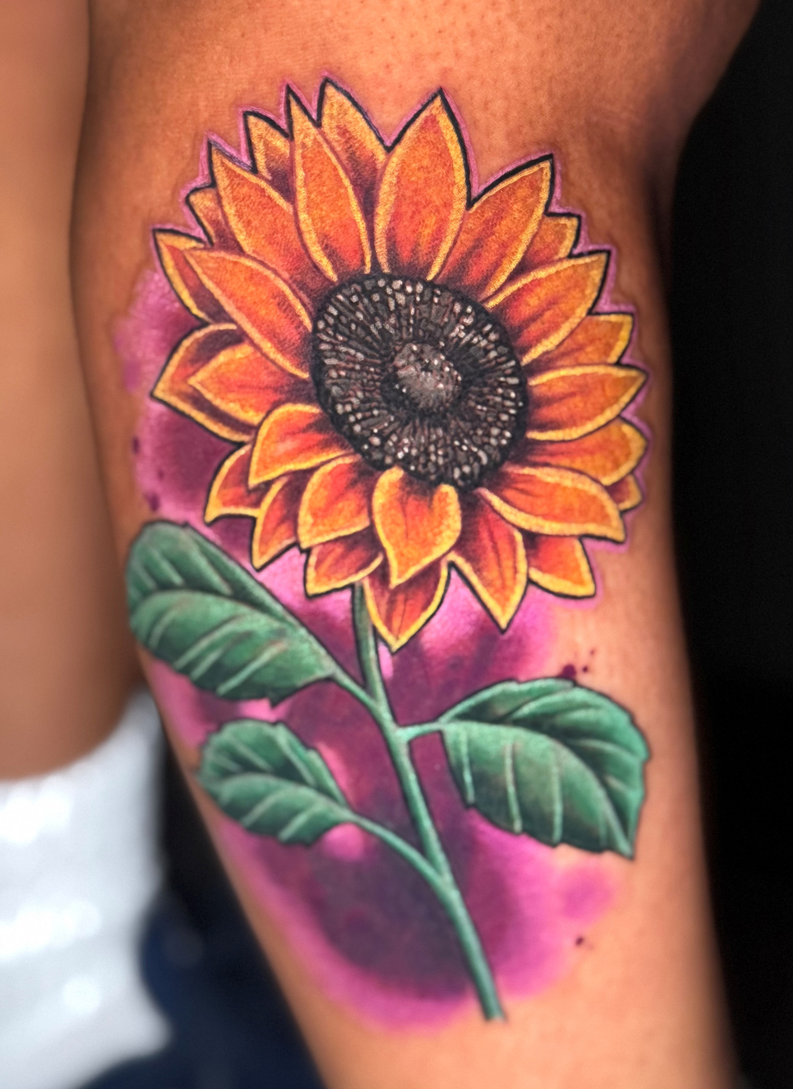 Sunflower tattoo with orange petals, black center, green leaves, and purple background.