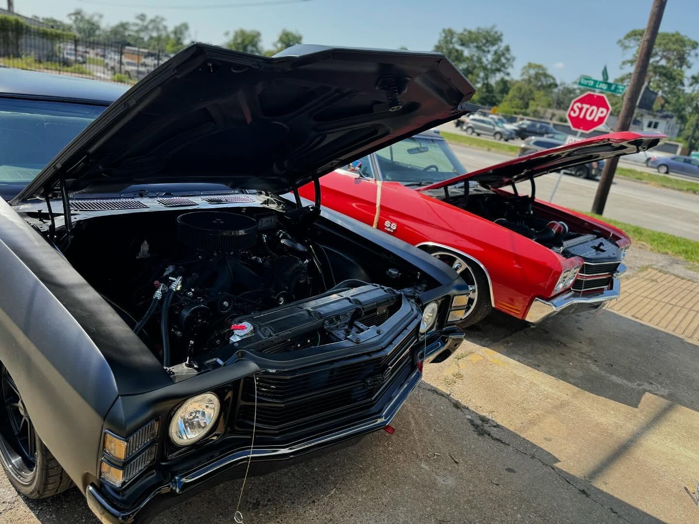 Two classic cars with open hoods, one black and one red, parked on a street near a stop sign.