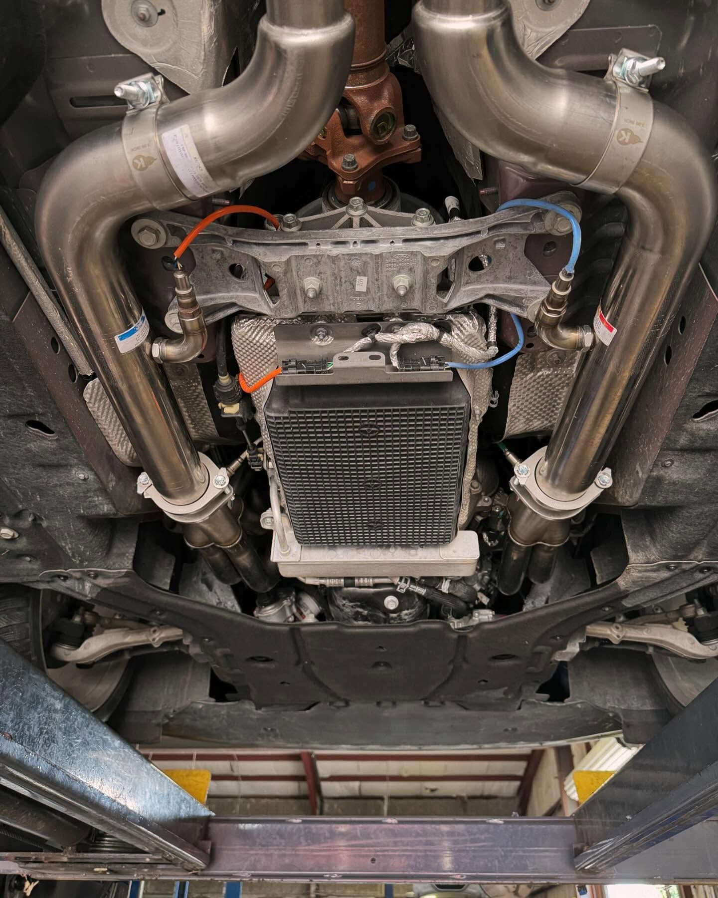 Undercarriage of a vehicle on a lift, showing exhaust pipes, heat shields, and other mechanical components.