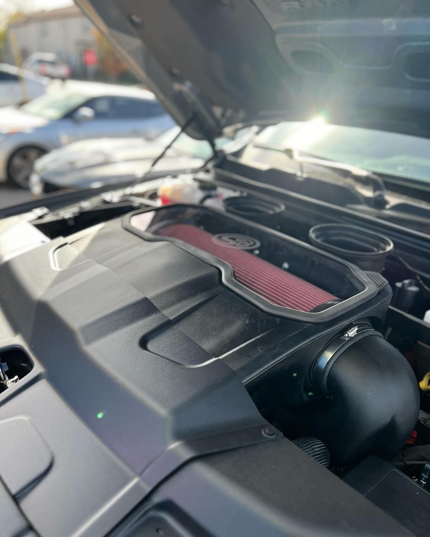 Close-up of a car engine bay, showing a black intake system with a red air filter. Hood is open in daylight.