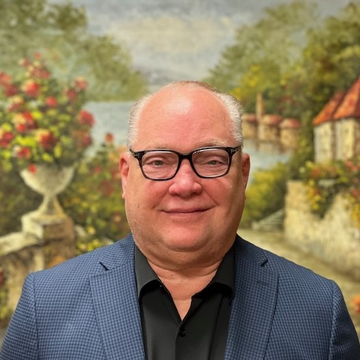 A man in a suit and glasses is smiling in front of a painting.