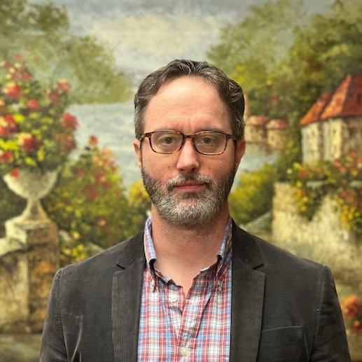 A man with a beard and glasses is standing in front of a painting.