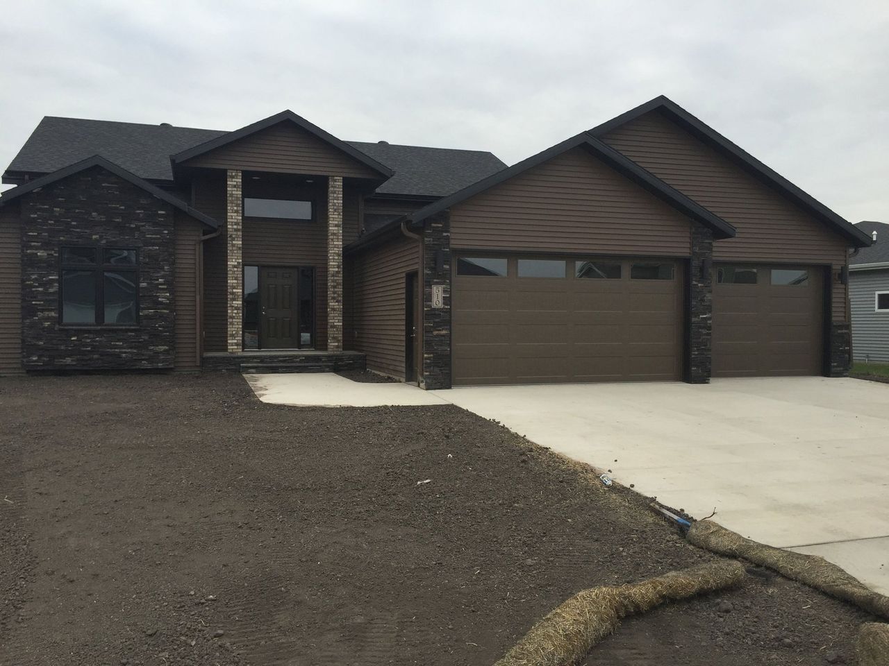 Suburban house with brick and wood exterior, two-car garage, and gravel driveway under overcast sky