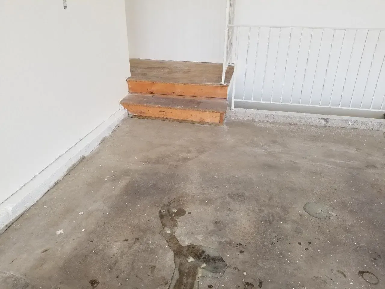 Concrete floor with dark stain and small wooden steps beside a white wall and railing.