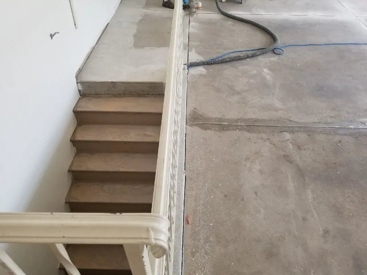 Indoor stairwell beside a concrete ramp, with a white railing and unfinished floor.