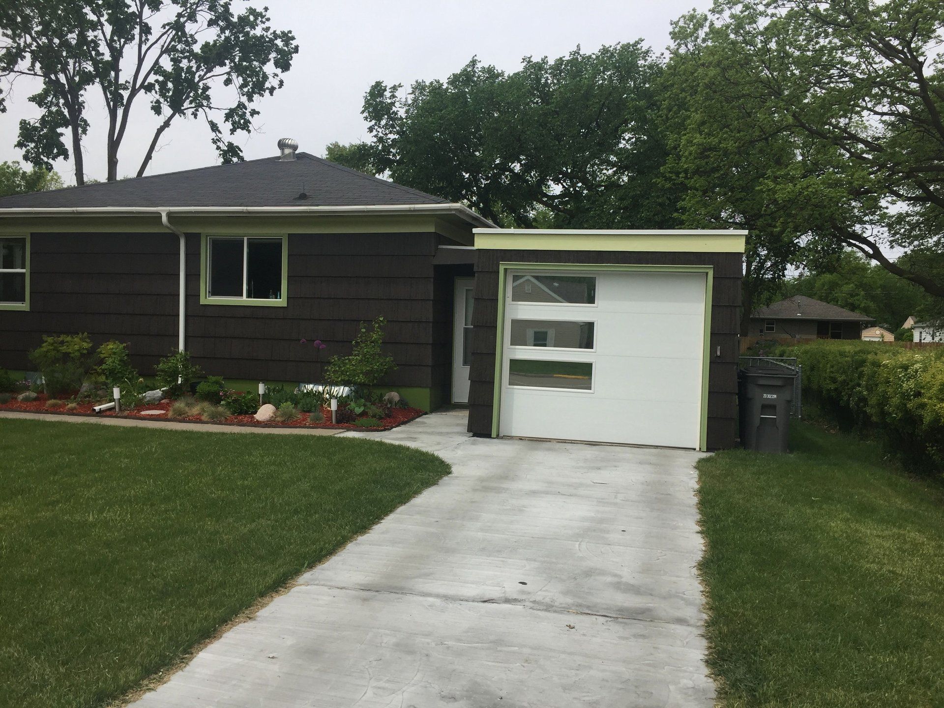 Small house with a white garage door and concrete driveway, surrounded by green lawn and trees.
