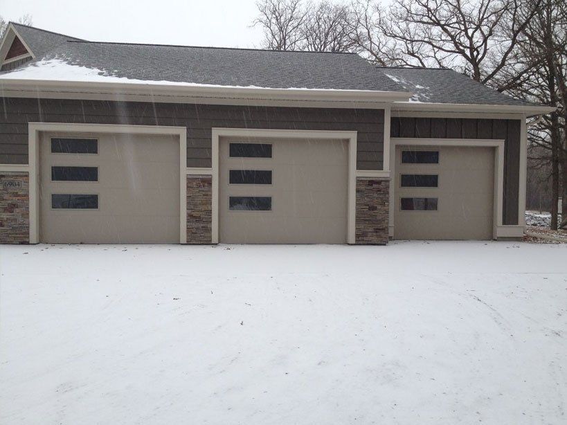 Snow-covered driveway in front of a three-car garage with stone accents and beige doors