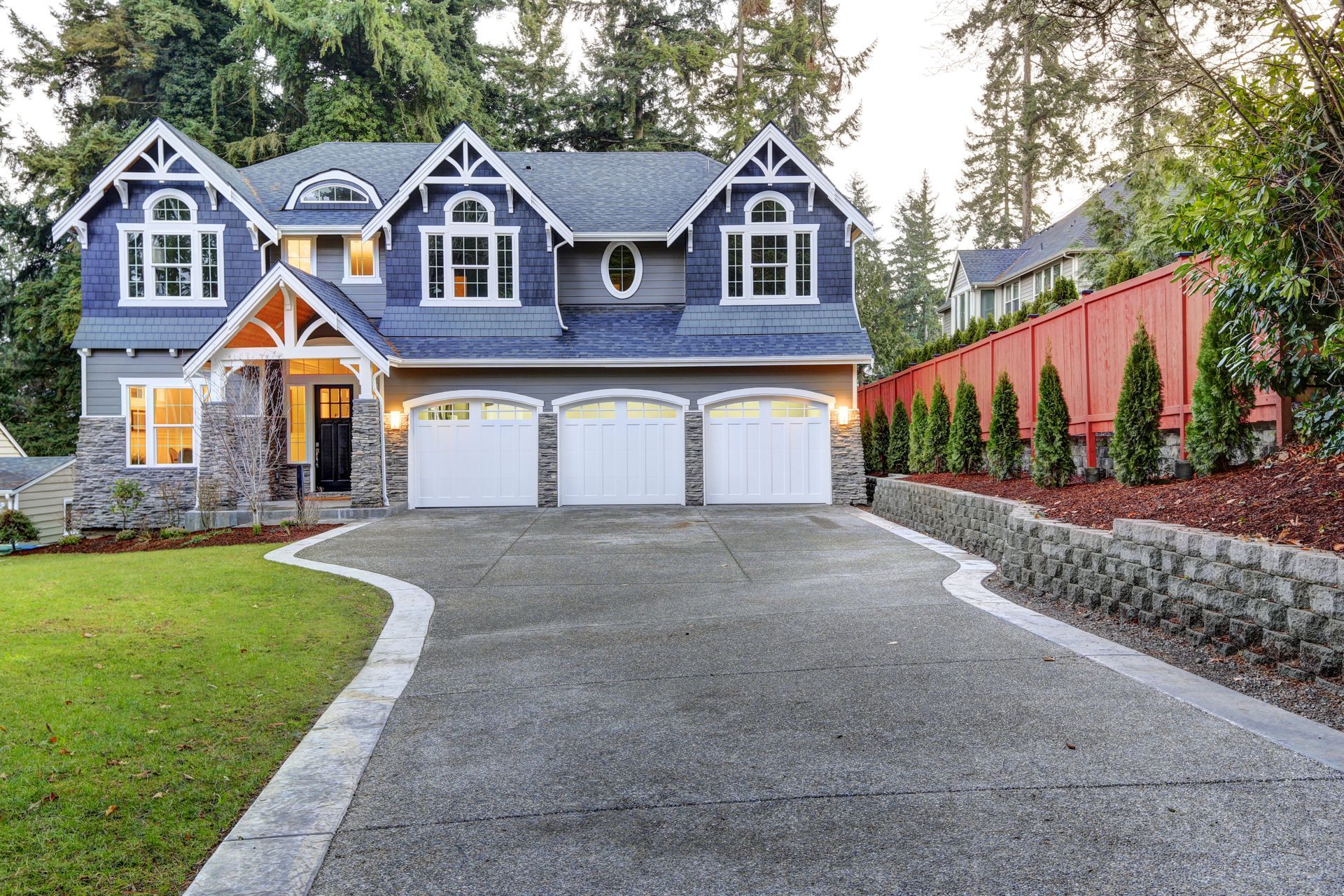 Large gray house with three-car garage, stone facade, and curved driveway surrounded by trees and hedges