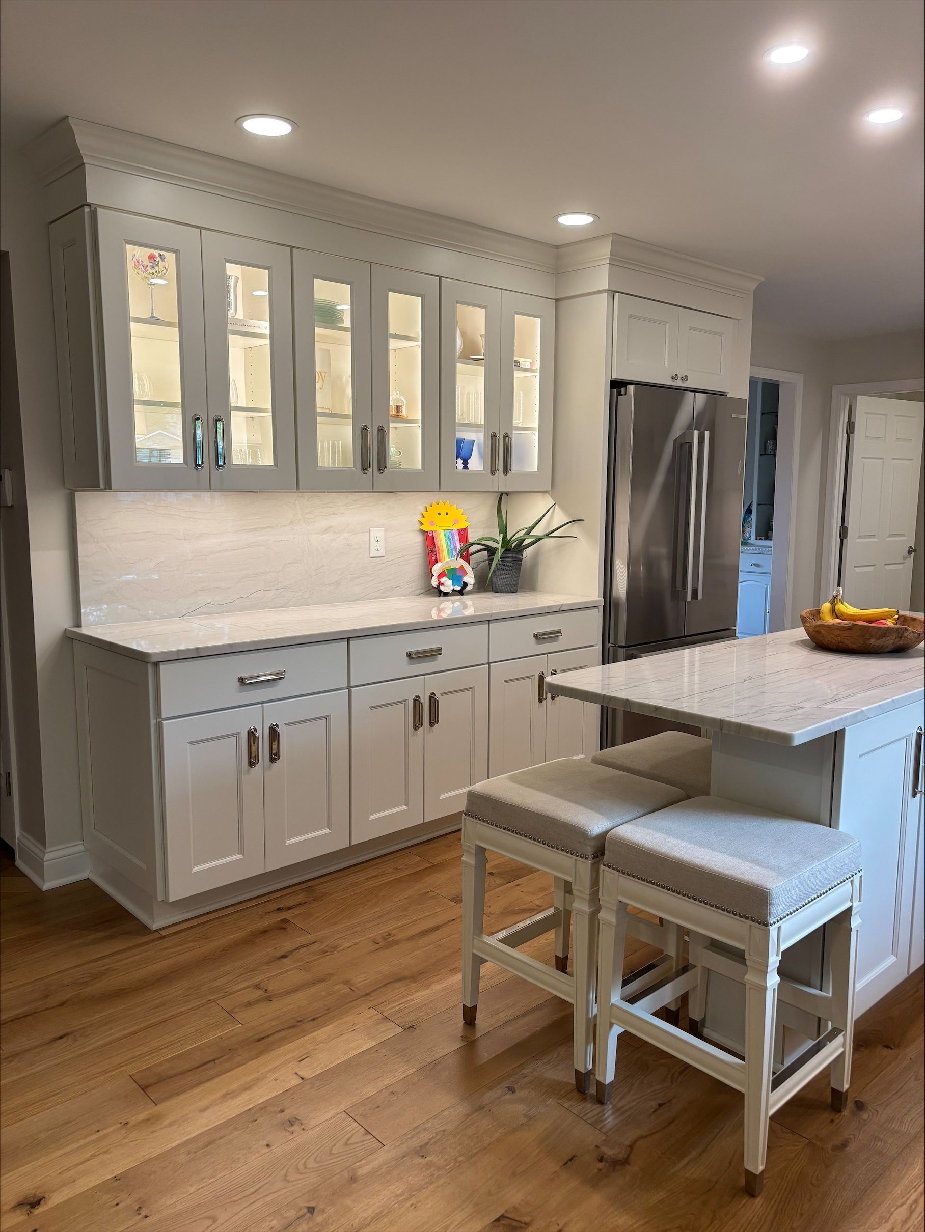 A kitchen with white cabinets , granite counter tops , stools and a refrigerator.