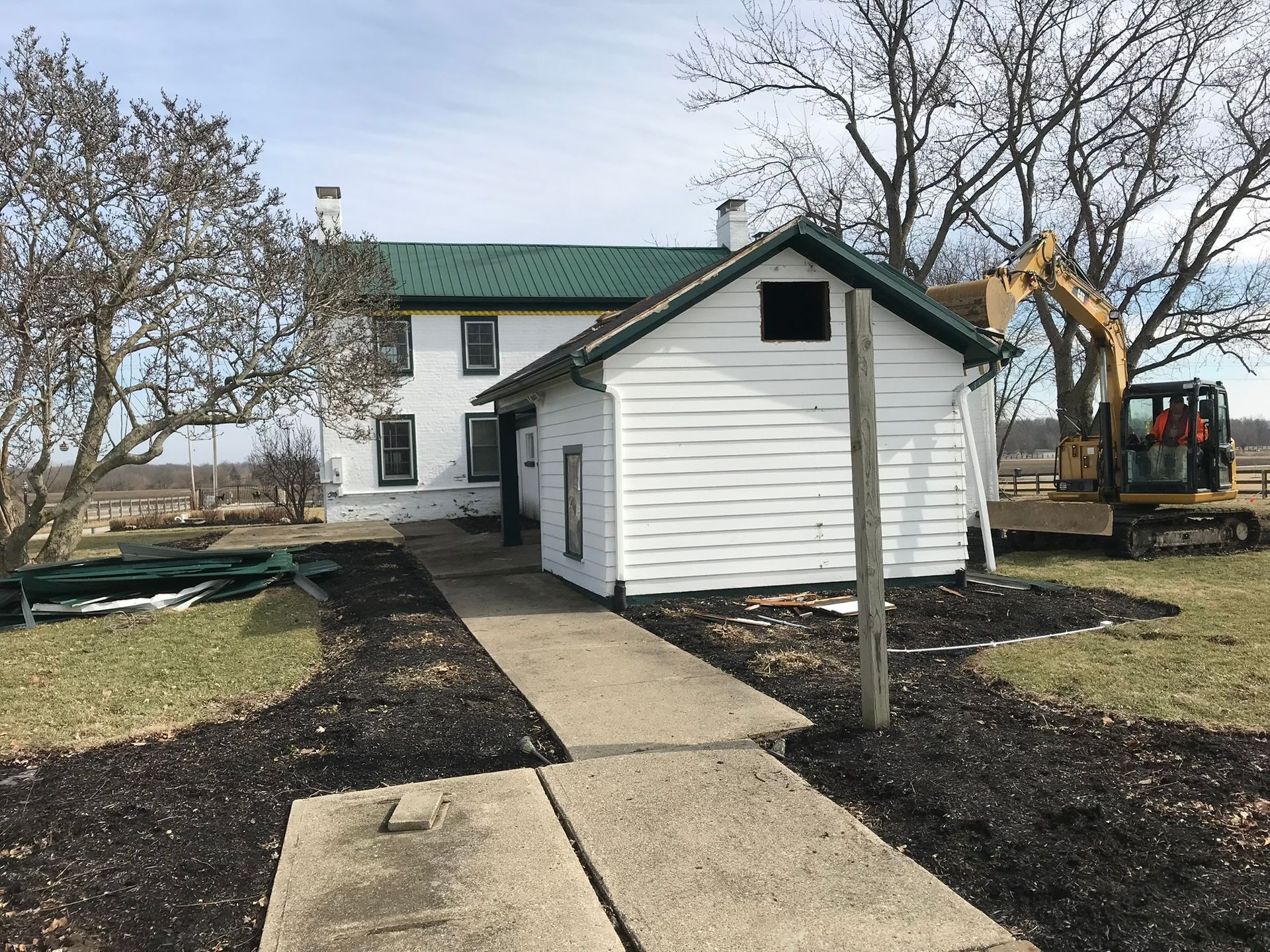 White house with green roof and side shed, concrete walkway, and excavator in a yard with bare trees.