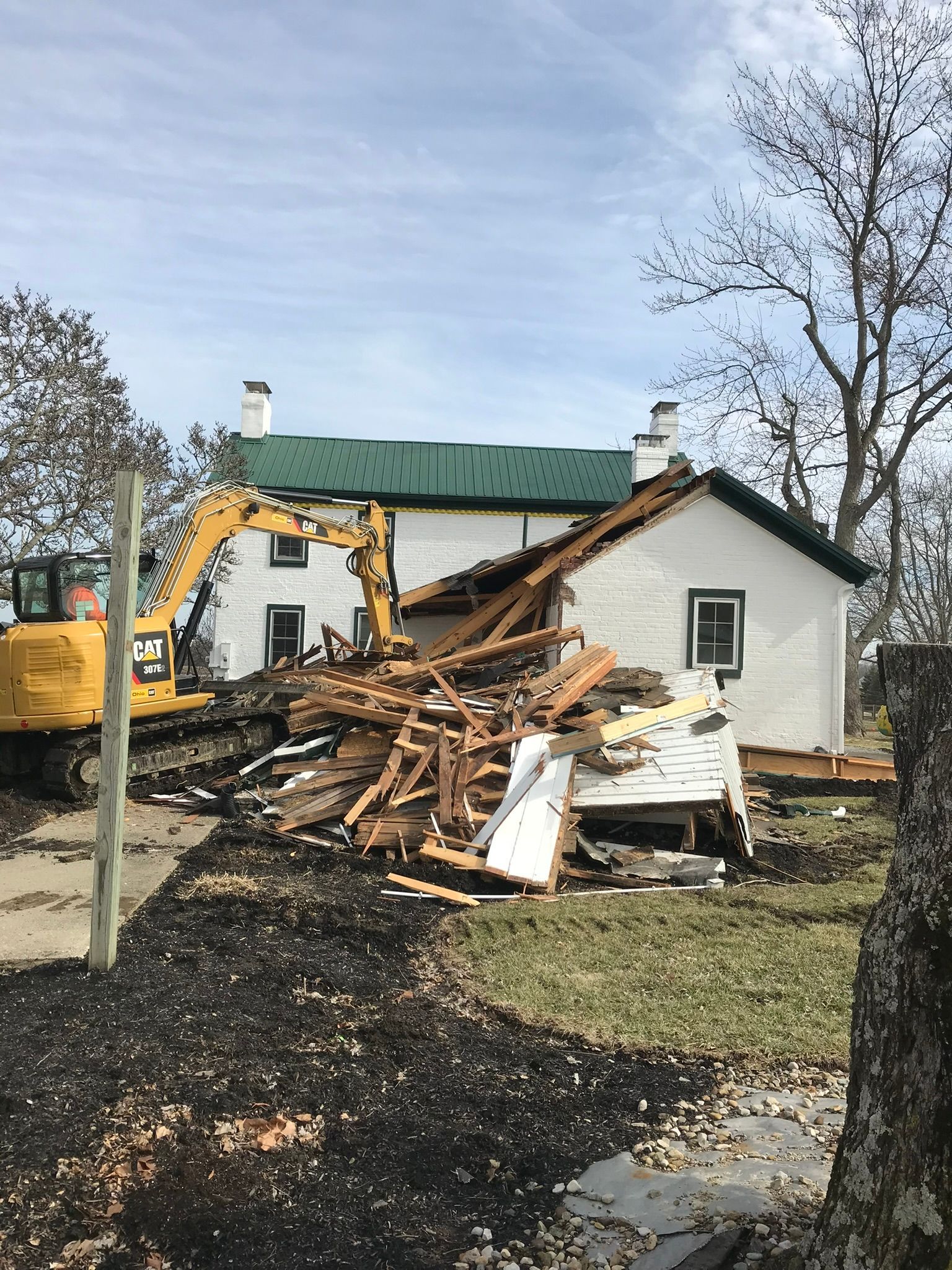 Excavator demolishing a damaged house with debris piled in front, under a cloudy sky.