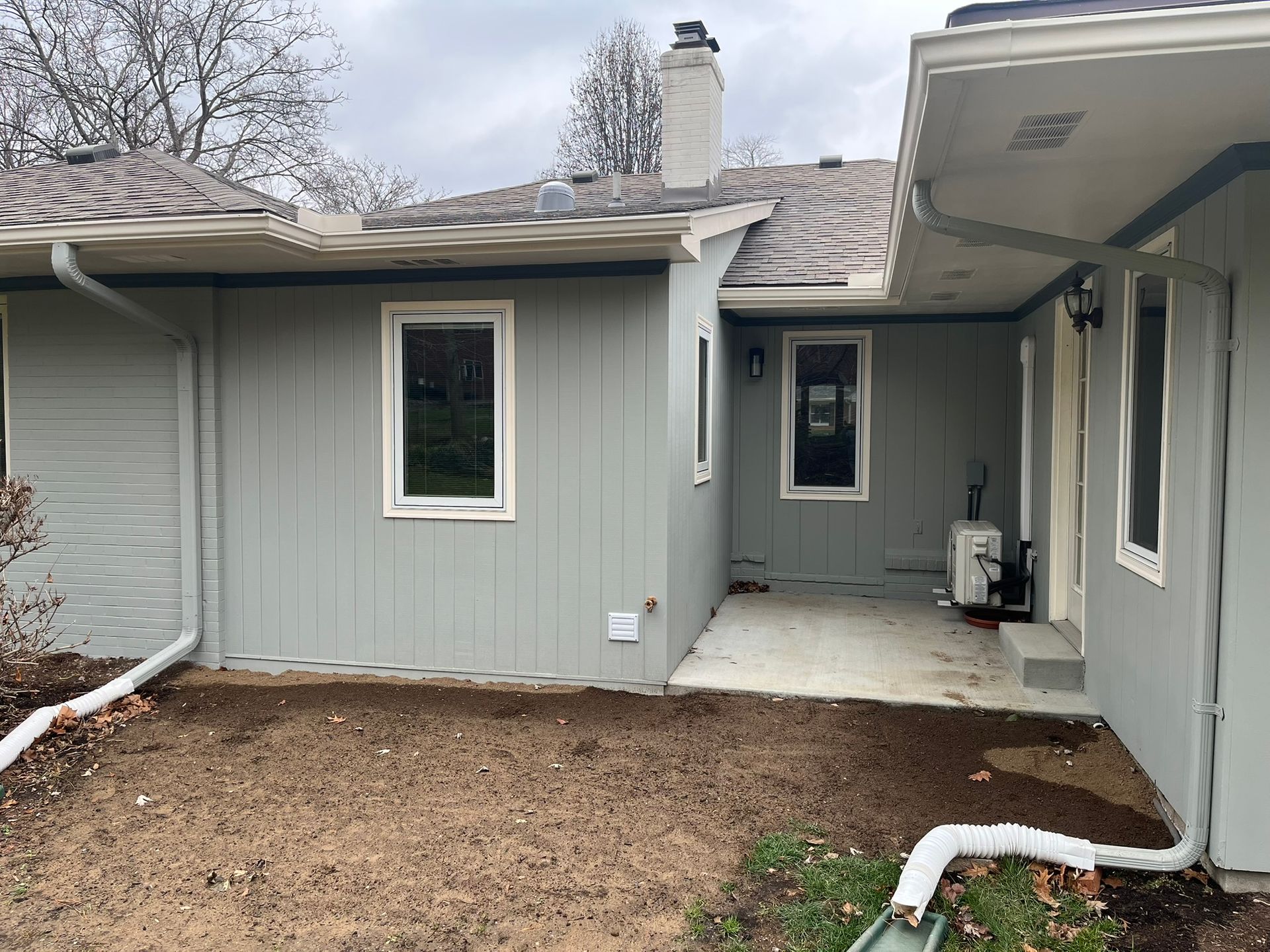 Gray house exterior with small patio, windows, downspouts, and bare backyard soil