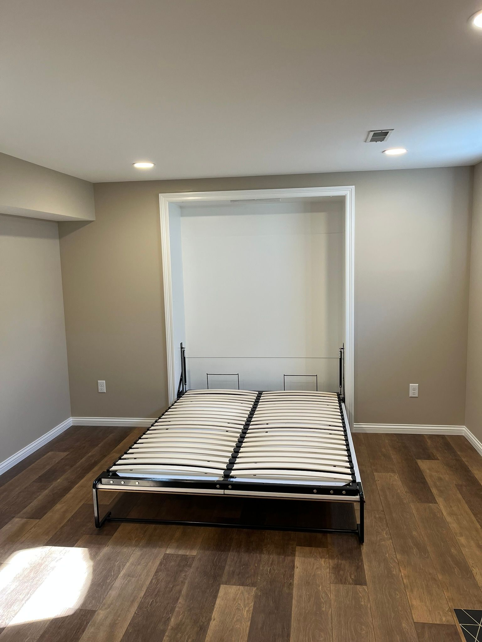Empty bedroom with a bed centered beneath a window in a beige room with wood floors.