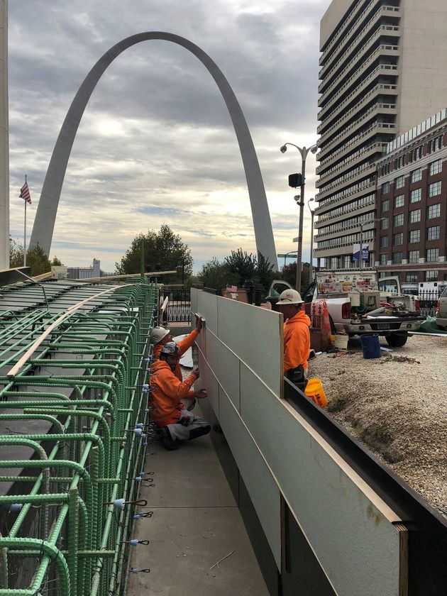 Workers in orange vests repairing a street near the Gateway Arch on a cloudy day.