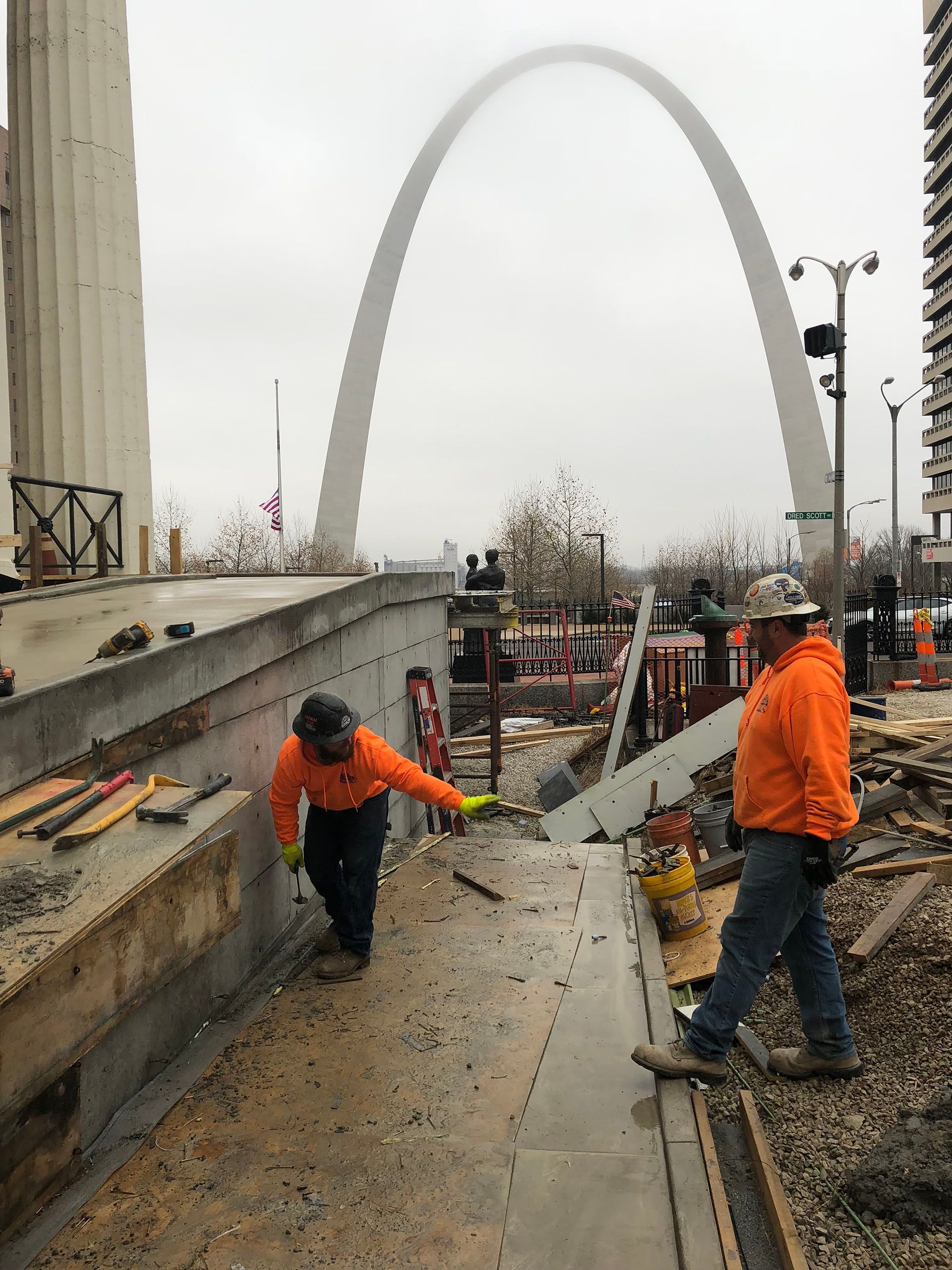 Workers in orange jackets on a construction site with the Gateway Arch in the background