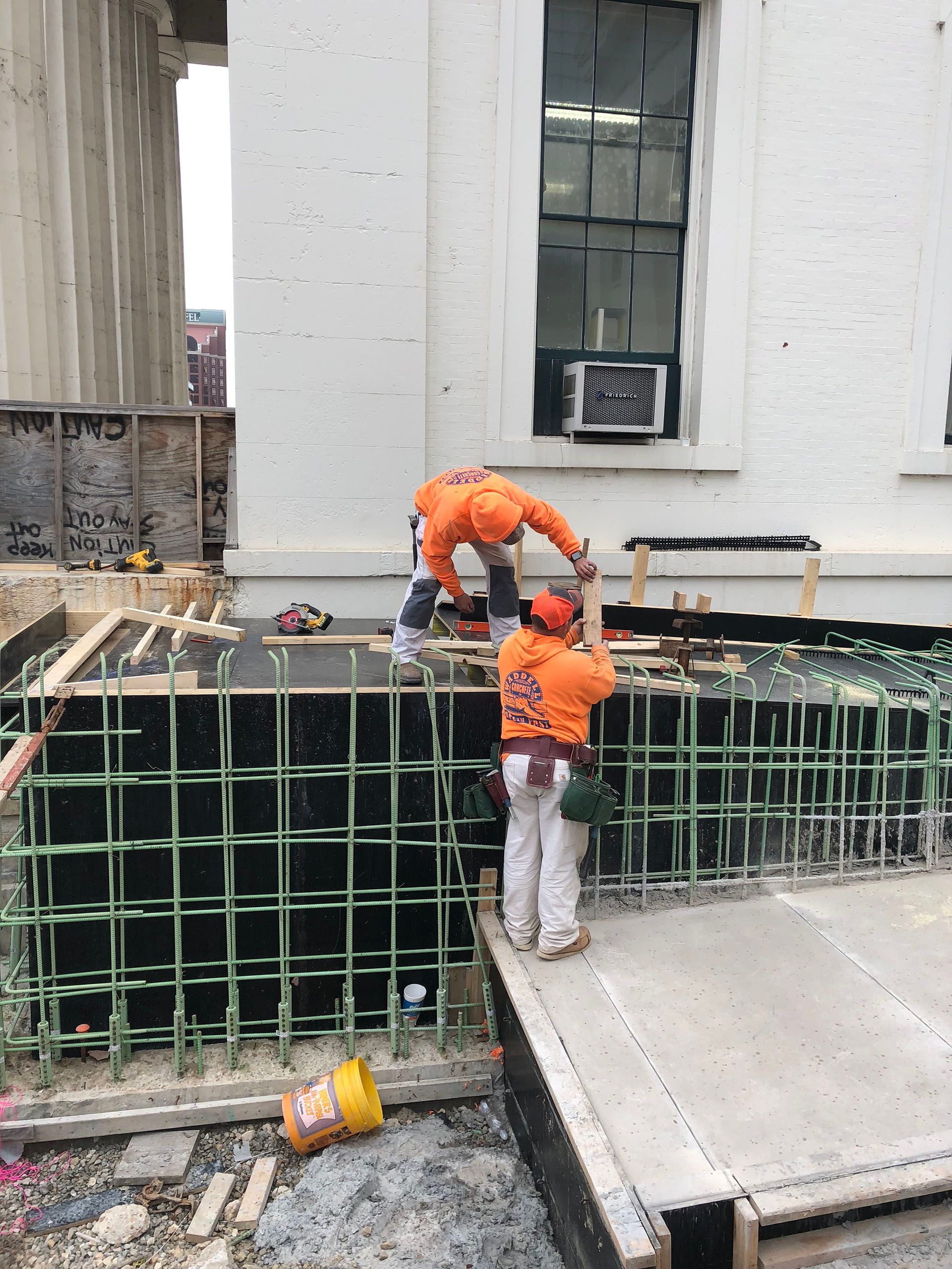 Workers in orange safety gear install rebar and formwork at a construction site.