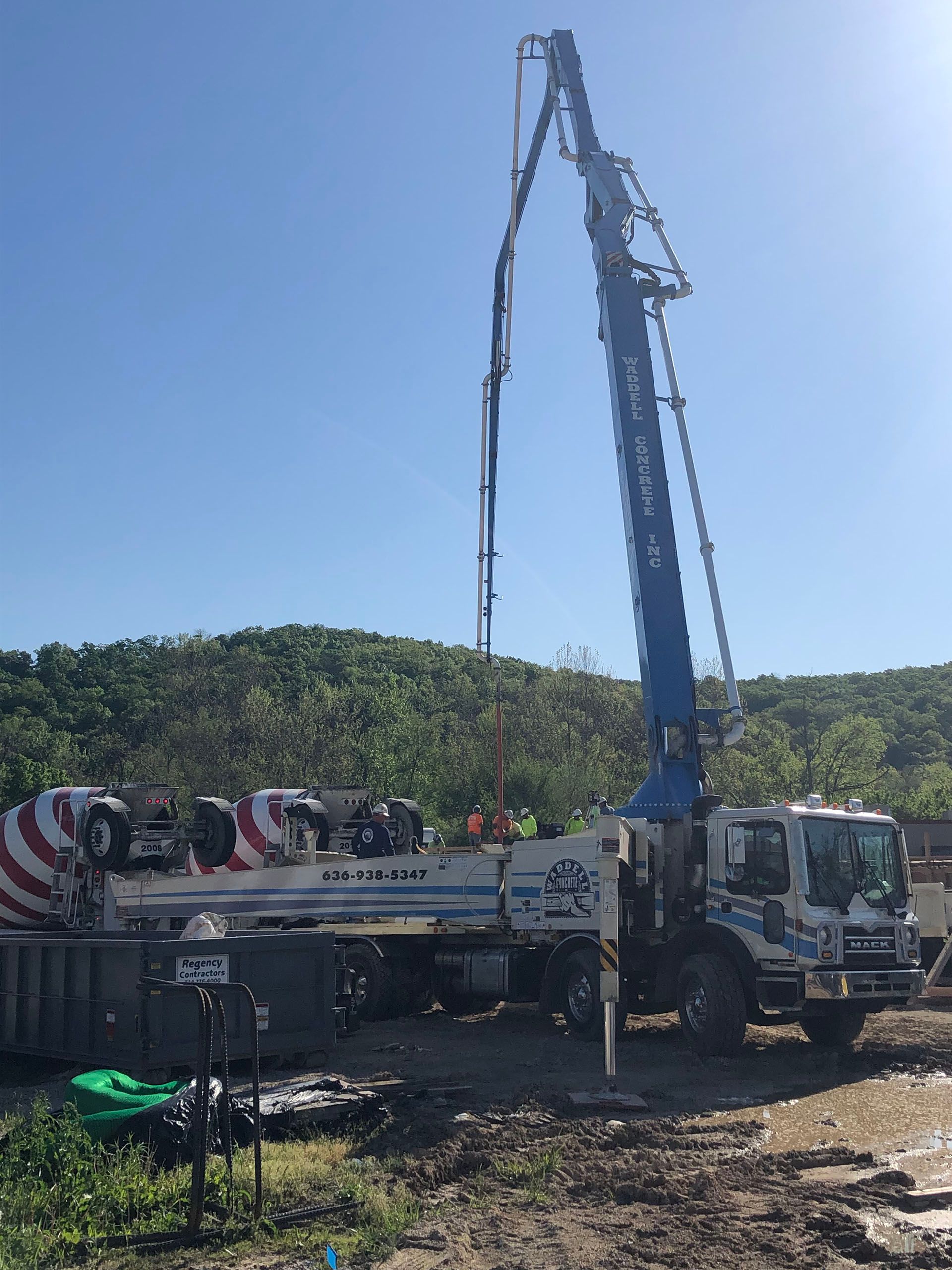 Blue crane truck lifting material from a flatbed in a sunny outdoor worksite near trees.