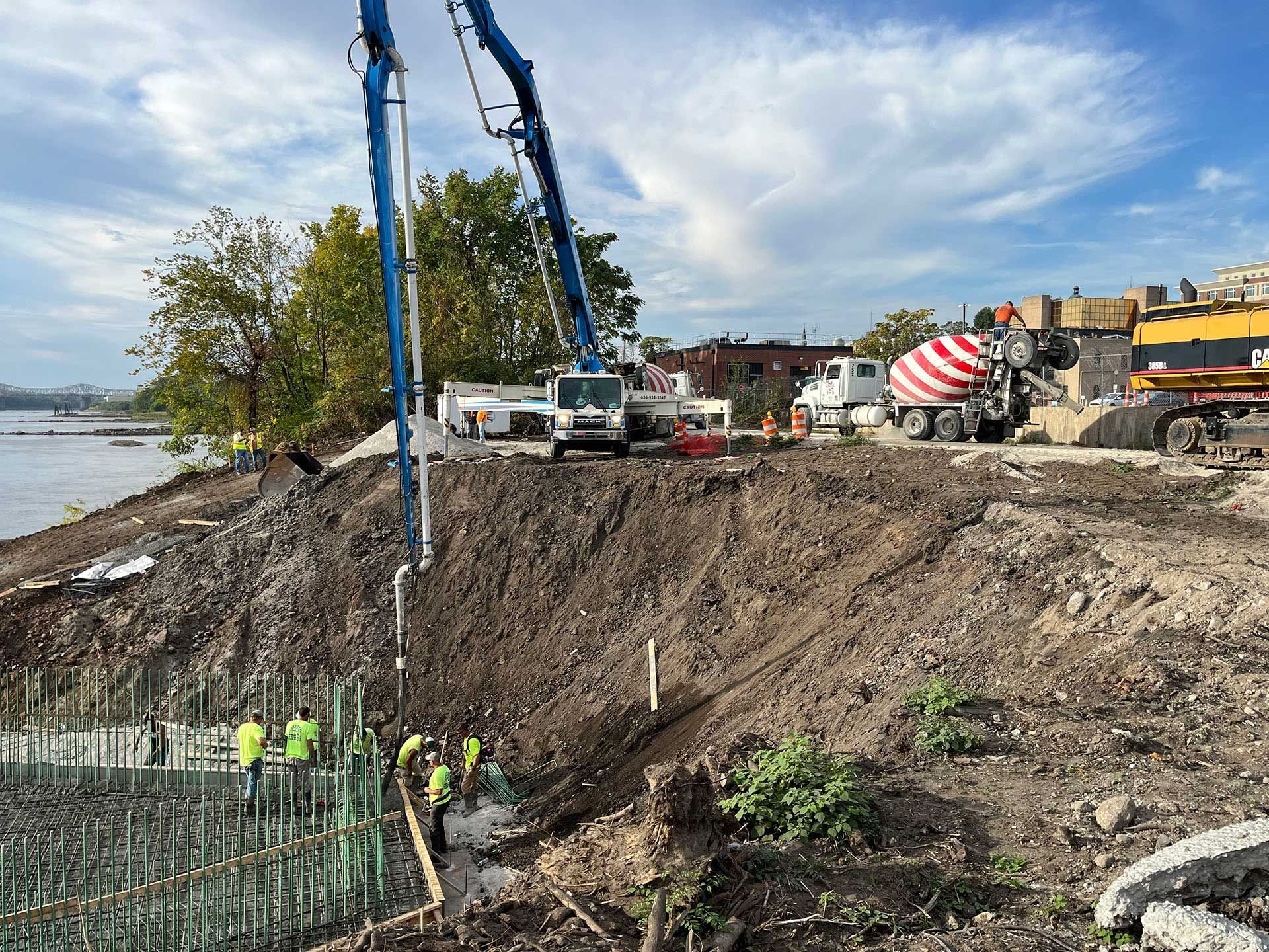 Construction site on a muddy riverbank with concrete pumps, trucks, and workers near the water