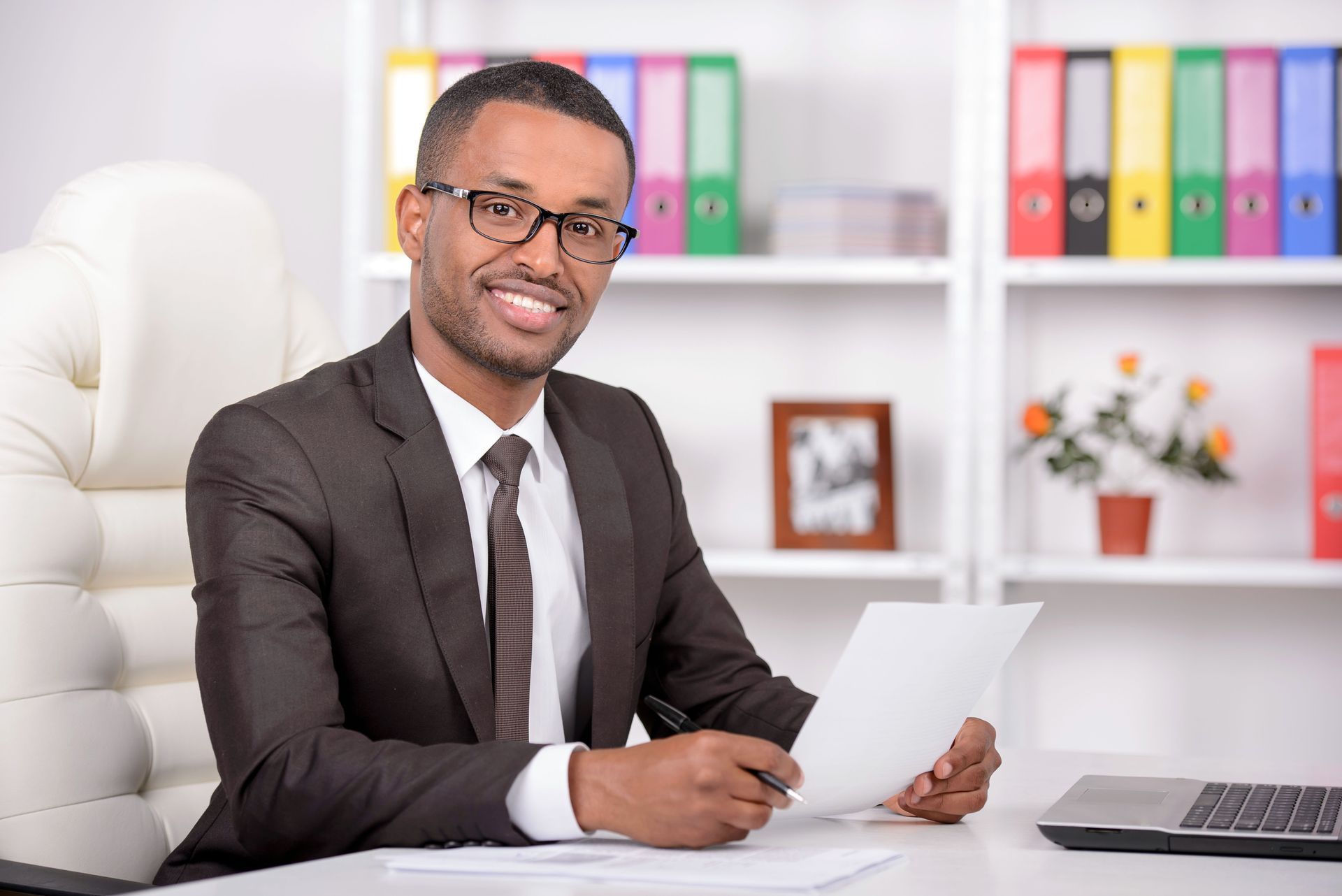 Office worker in a suit smiling at a desk with papers, laptop, and colorful binders on shelves behind him